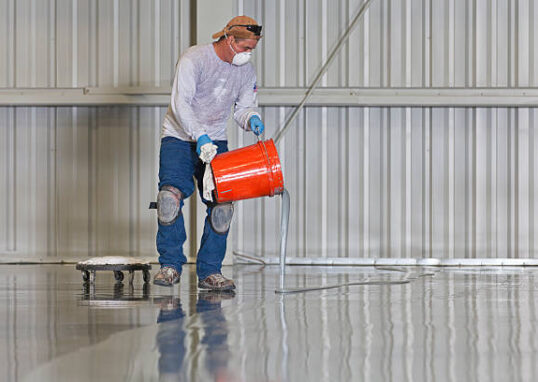 A construction worker pours paint onto a warehouse floor.